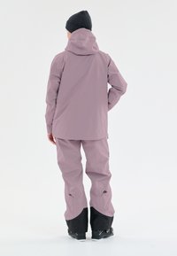 Vedenkestävä hiihtotakki ja housut vaaleanpunaisena. Takissa on huppu ja pidempi malli; housuissa on mustia yksityiskohtia ja säädettävät hihansuut.