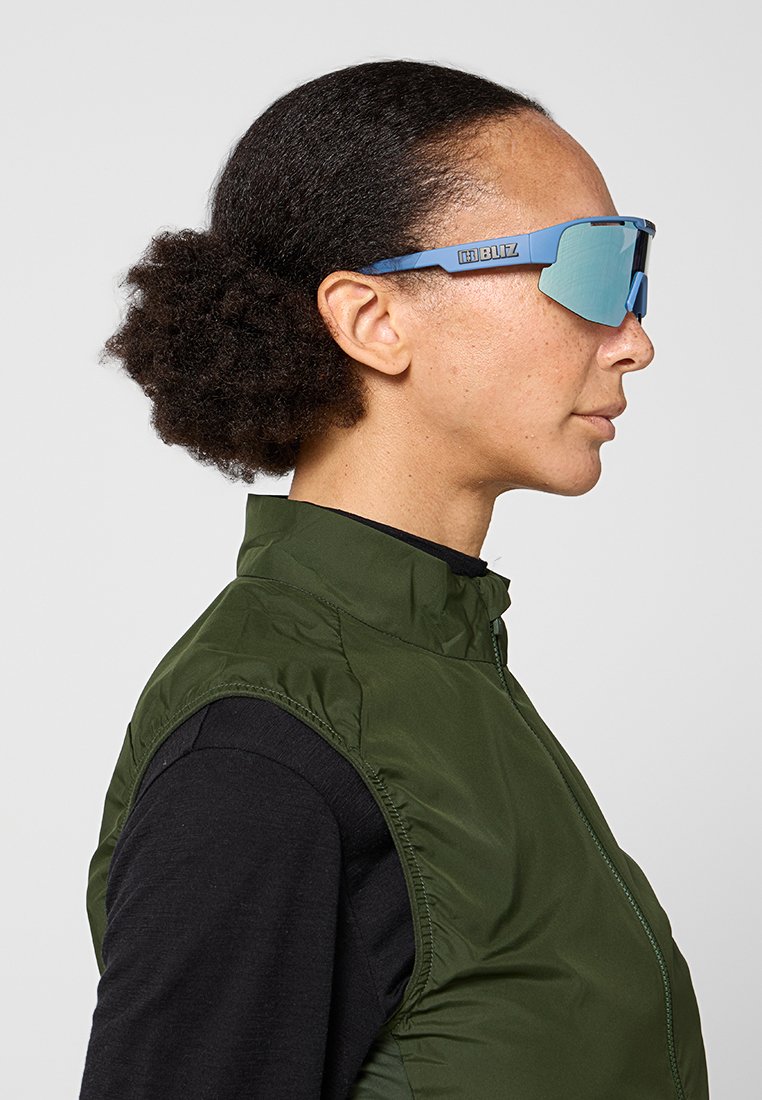 Sonnenbrille mit blauen Rahmen und reflektierenden Gläsern, mit einem schlanken, modernen Design. Die Person trägt eine grüne Weste über einem schwarzen Shirt.