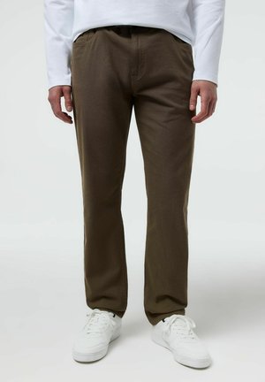 Mężczyzna ubrany w brązowe spodnie o prostych nogawkach, białą koszulę z długim rękawem i białe niskie trampki, stojący na jasnoszarym tle.