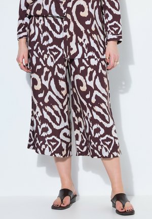 Wijde, korte broek met een paars en beige patroon, gecombineerd met een bijpassende top met lange mouwen, zwarte sandalen met open tenen en hakken, en gouden ringen aan de vingers.