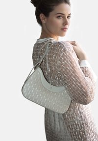 Cremefarbener Lederhandtasche mit geschwungenem Design, verziert mit einem strukturierten Blattmuster und goldenen Beschlägen. Das Modell trägt ein durchscheinendes Oberteil mit detaillierter Stickerei.