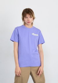 Purple short-sleeve t-shirt made of cotton, featuring a white logo on the left chest, paired with beige pants. Soft texture, casual design.