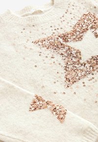 Pull tricoté crème avec une grande étoile en sequins or rose sur le devant et un petit détail étoile sur le poignet de la manche.