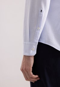 Camicia bianca a maniche lunghe con colletto dritto, dotata di bottoni sui polsini e un motivo di rosa nera ricamata vicino alla manica.