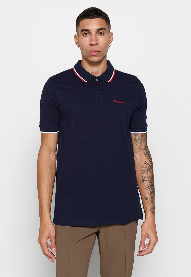 Polo shirt navy en tissu texturé, avec un col et des poignets rayés rouge et blanc, logo brodé sur la poitrine, manches courtes.