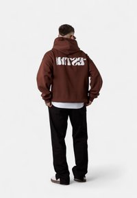 Sweat à capuche marron avec un grand texte blanc au dos. Coupe décontractée, capuche à cordon et poignets côtelés. Porté par-dessus une chemise blanche et un pantalon noir.