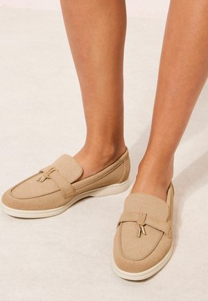 Hellbraune Slipper-Loafer mit weißen Sohlen und kleinen goldfarbenen Quasten, barfuß getragen vor hellem Hintergrund.