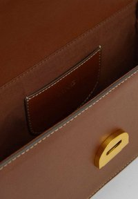 Interno di una borsa in pelle marrone con una texture liscia, che presenta un dettaglio in metallo dorato e una tasca laterale per l'organizzazione.