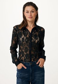 Zwarte kantblouse met bloemenpatroon, knoopsluiting aan de voorkant en zwarte kraag; doorzichtige stof en lange mouwen; gecombineerd met blauwe denim jeans.