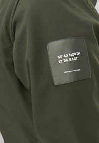Grøn sweatshirt med et sort label på venstre ærme med hvid tekst: "55° 40' NORD 12° 36' ØST JACK & JONES ©1990."