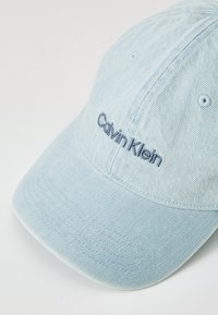 Light blue denim baseball cap with embroidered "Calvin Klein" logo in dark blue. Features stitching details and metal ventilation eyelets.