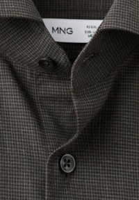 Camisa abotonada con un patrón de cuadros marrón oscuro, que presenta un cuello clásico y un botón negro. La tela tiene una textura sutil.