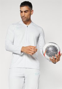 Hvit treningsjakke med glidelås foran og Nike-logo i turkis. Modellen holder en mønstrete fotball og har på seg matchende hvite bukser.