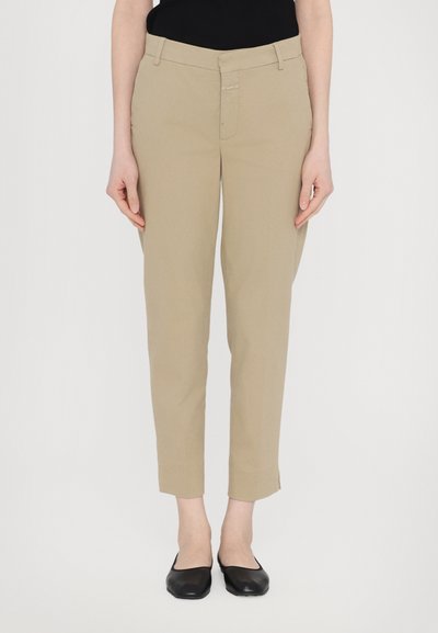 Pantalones beige ajustados con un corte entallado, que presentan un cierre delantero con cremallera y botón, y dobladillo a la altura del tobillo. Combinados con zapatos planos negros.