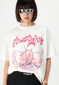 White oversized t-shirt featuring a pink graphic design of a sun and mountain. Black logo waistband visible below, paired with sunglasses.