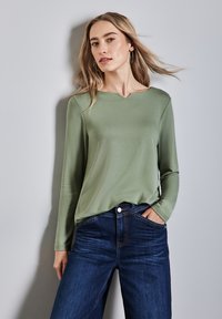 Maglietta a maniche lunghe di colore verde oliva con scollo ampio, abbinata a jeans denim blu scuro. Texture liscia senza motivi o hardware visibili.