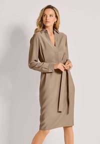 Beige jurk met lange mouwen en een kraag, met een v-hals, zijzakken en een bijpassende riem in de taille. Gladde textuur.