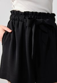 Pantalones cortos negros de tejido suave que cuentan con una cinturilla elástica con volantes, bolsillos laterales y un cinturón de tela a juego atado en la parte delantera.