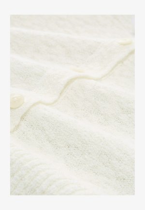 Cremefarbener, strukturiert gestrickter Stoff mit geripptem Rand und durchsichtigen Knöpfen, der einen Teil der mit Knöpfen versehenen Vorderseite eines Kleidungsstücks zeigt.