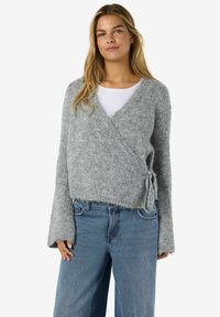 Kvinna som bär en ljusgrå, fluffig kappa knuten i midjan över en vit topp och lösa blå jeans, stående mot en enkel bakgrund.