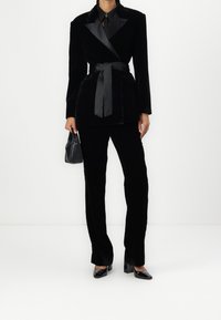 Czarny aksamitny garnitur o dopasowanym kroju, z kurtką z paskiem, satynowym kołnierzem i pasującymi prostymi spodniami. Towarzyszy mu mała czarna torebka.