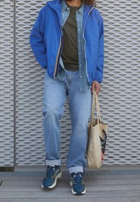 Blauw lichtgewicht jack met witte accenten over een groen shirt en een denim overhemd. Lichtblauwe jeans en blauwgroene sneakers. Beige boodschappentas.