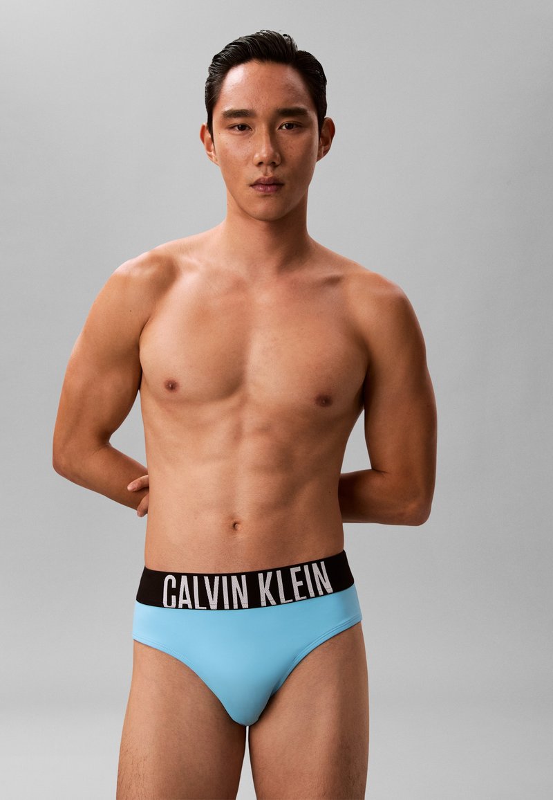 Mladý muž stojí s rukami za chrbtom, oblečený v svetlomodrých slipoch Calvin Klein s čiernym pásikom na jednoduchom sivom pozadí.