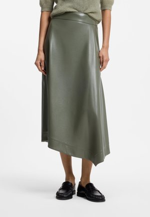 Kvinde iført grøn asymmetrisk midi-nederdel, beige striktrøje og sorte læderloafers står på en ensfarvet hvid baggrund.