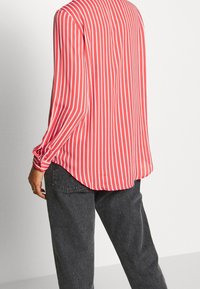 Red and white striped shirt with long sleeves, buttoned cuffs, and a rounded hem, worn with dark gray pants. Soft fabric, relaxed fit.