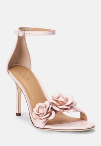 Różowe sandały na wysokim obcasie z paskiem na kostkę, z dwoma dekoracyjnymi kwiatami z tkaniny z przodu i gładkim, skórzopodobnym wykończeniem.