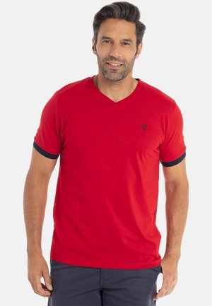 Homme aux cheveux noirs courts et à la barbe, portant un T-shirt rouge à col en V avec des bordures bleu marine sur les manches et un pantalon sombre, se tenant devant un fond blanc.
