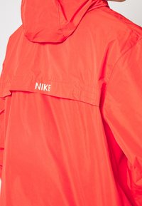 Röd Nike vindjacka med huva, med en slät textur, elastiska ärmslut och en ventil bak. Logotyp i vitt tryckt på den övre ryggen.