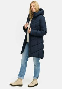 Marineblauwe pufferjas met capuchon, geborduurd ontwerp, rits aan de voorkant en patchdetail. Gecombineerd met lichtblauwe jeans en crèmekleurige enkellaarsjes.