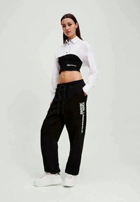 Czarny krótki top z białą koszulą z długim rękawem, w połączeniu z czarnymi spodniami dresowymi z białym logo. Model ma na sobie białe trampki.