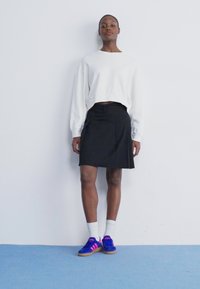 Λευκό κοντό φούτερ, μαύρη φούστα, μοβ και ροζ αθλητικά παπούτσια με λευκές κάλτσες. Το μοντέλο στέκεται μπροστά από έναν απλό τοίχο, επιδεικνύοντας την εμφάνιση.