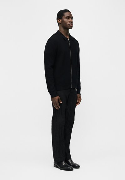 Άντρας που στέκεται φορώντας μαύρο πλεκτό πουλόβερ με φερμουάρ, υφασμένα μαύρα παντελόνια και μαύρα δερμάτινα παπούτσια σε φόντο απλό λευκό.