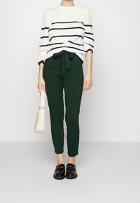 Biały sweter z dzianiny w granatowe paski, zielone spodnie z wiązaniem w pasie oraz czarne loafersy, w zestawieniu z kremową torbą na ramię.