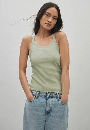 Kvinde med langt mørkt hår iført en salviegrøn tanktop og lyseblå jeans, stående med hænderne i lommerne imod en ensfarvet baggrund.