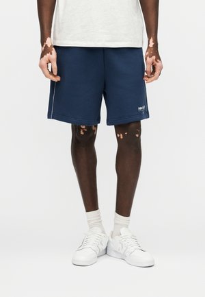 Person med vitiligo iført marineblå shorts, hvide sokker og hvide sneakers, stående mod en ensartet lys baggrund.