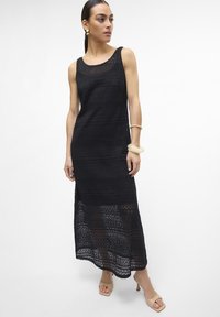 Vestido maxi de crochê preto com decote arredondado, apresentando um padrão texturizado e seções transparentes na barra. Combinado com sapatos de salto nude.