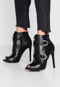 Czarne botki na wysokim obcasie z odkrytymi palcami, wykonane z gładkiej skóry, z dekoracyjnymi paskami i detalami z srebrnym zamkiem.