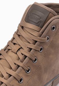 Zapato alto marrón con material sintético texturizado, ojetes de metal y cordones planos de tejido. Presenta un parche con logo en la lengüeta.