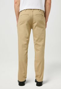 Beige Chinos mit geradem Bein, ausgestattet mit zwei Gesäßtaschen, die feine Nähdetails aufweisen, und einem glatten Baumwollmischgewebe.