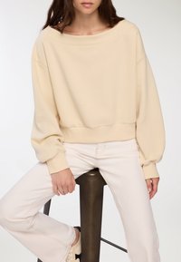 Sweatshirt beige a cropped con scollatura ampia, spalle cadenti e polsini a coste. Abbinato a pantaloni chiari e seduto su uno sgabello di metallo.