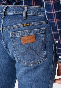 Jeans de ganga azul com um patch de pele da Wrangler, costura em contraste e um bolso traseiro com um distinto design em chevron.