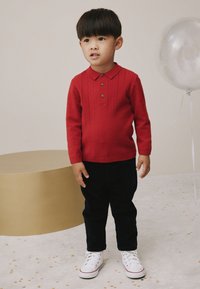 Rood gebreid poloshirt met kraag en drie knopen, gecombineerd met donkerblauwe broek en witte sneakers. Gouden ronde tafel op de achtergrond.