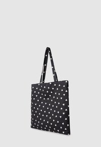 Czarna torba tote z kremowymi wzorami serc, płaskiego prostokątnego kształtu, z dwoma długimi uchwytami do noszenia w ręku lub na ramieniu.