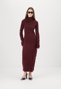 Burgunderfarbnes Strickkleid mit hohem Kragen, seitlichen Knöpfen, langen Ärmeln und seitlichen Schlitzen; kombiniert mit schwarzen High Heels und Sonnenbrille.