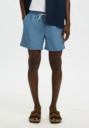 Mand iført midt-lår blå snørede shorts, mørkeblå skjorte, hvid top og brune sandaler med to stropper står mod en lys baggrund.