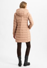 Pufferjacke in hellbeige mit Kapuze, ausgestattet mit horizontal gestepptem Design, kombiniert mit braunen figurbetonten Hosen und schwarzen Knöchelstiefeln.